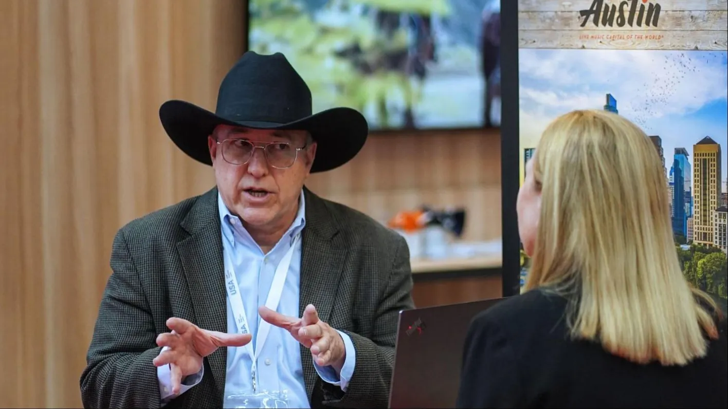 Man in cowboy hat engaging with an attendee, using expressive hand gestures during a booth talk