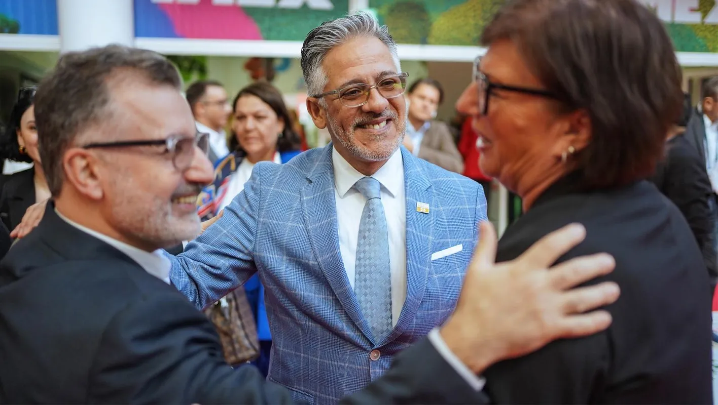 Two men and a woman, in suits, smiling and talking on IMEX Frankfurt's show floor.