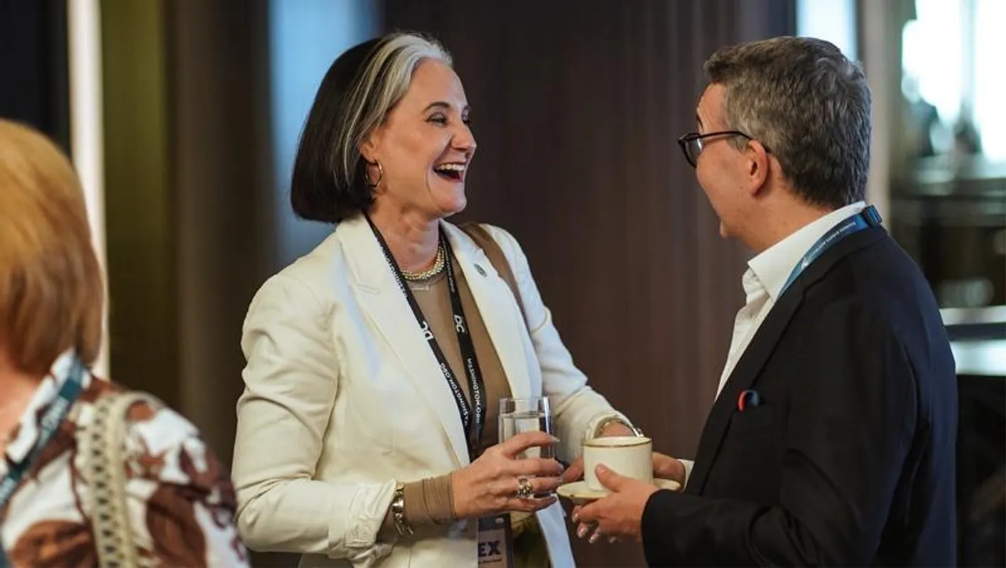 Two attendees with drinks, talking and laughing at IMEX Frankfurt. Both wearing suits and lanyards.
