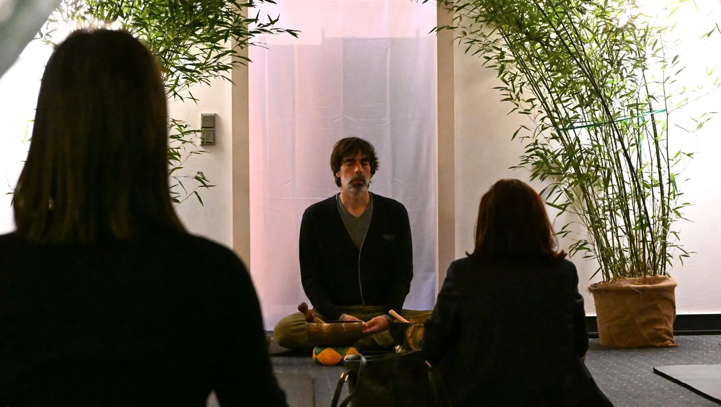 Person seated cross-legged holding a bowl, with two others in front, framed by bamboo.