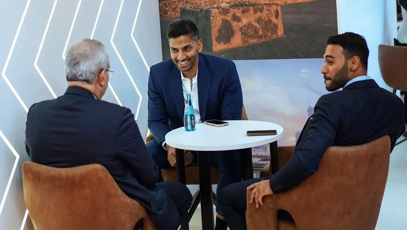Three men in suits in a meeting on IMEX Frankfurt's show floor. They're sat in brown chairs.
