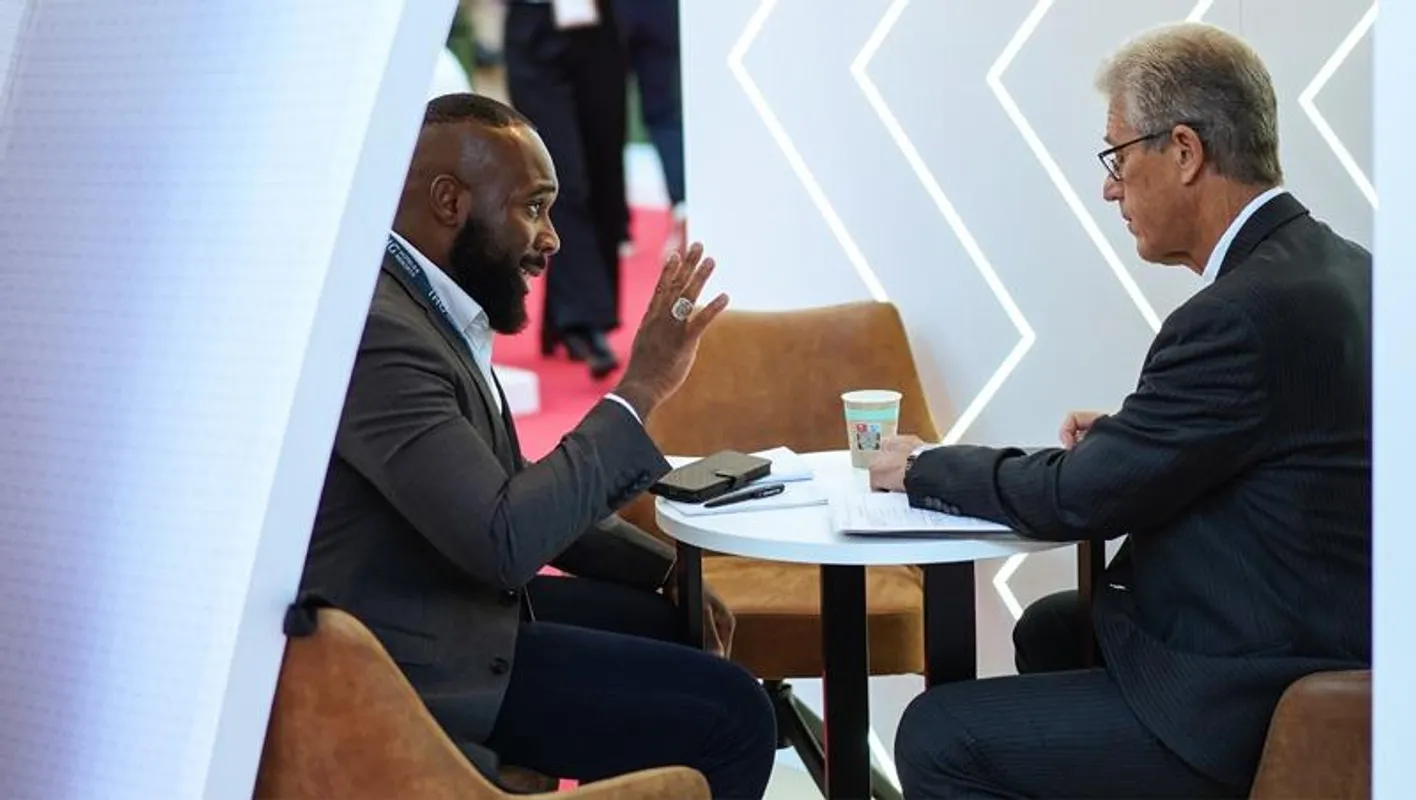 Two people in suits sit talking at a table in a white striped booth at IMEX Frankfurt. 