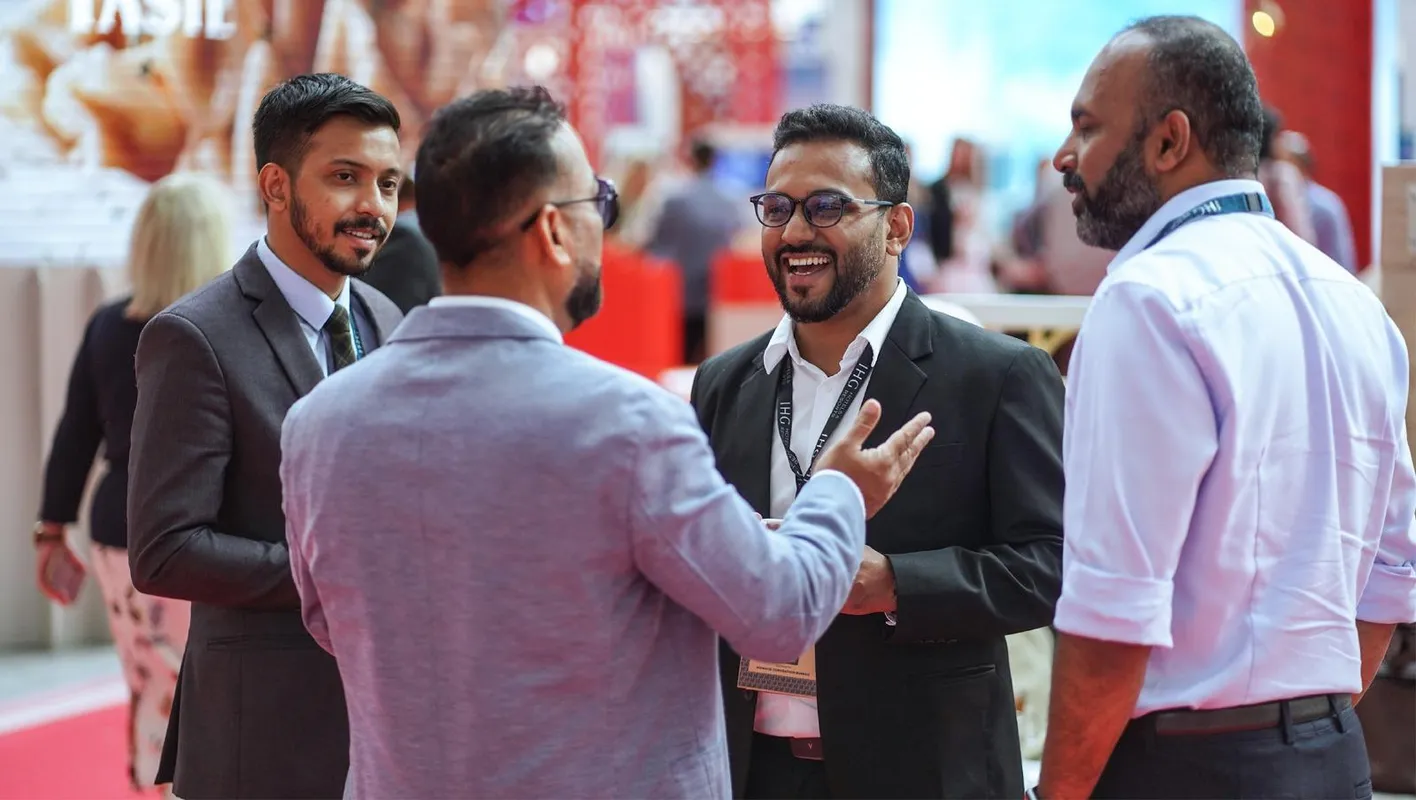 Four men stand in suits stand on the IMEX Frankfurt show floor talking and laughing.
