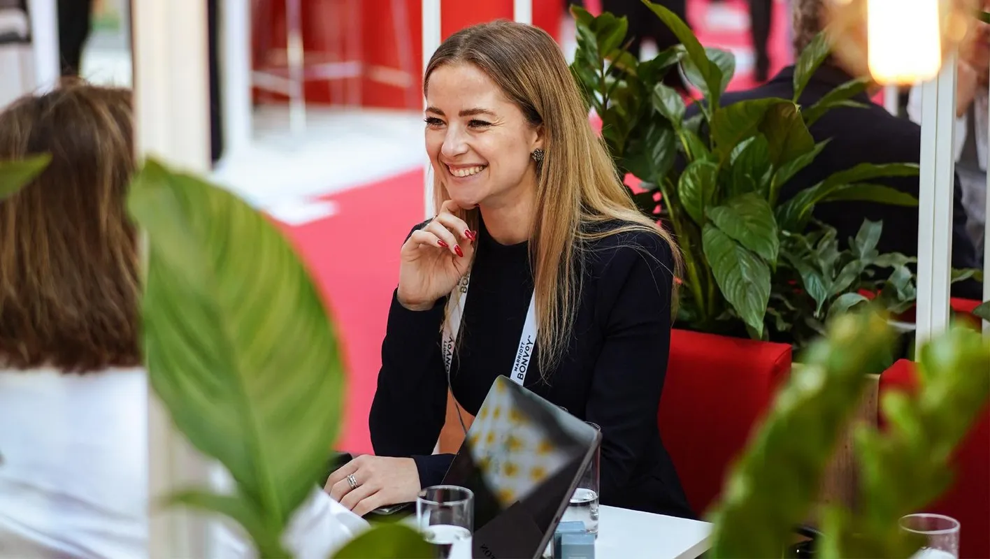 A woman sits smiling at a table in a red booth. She is surrounded by plants.