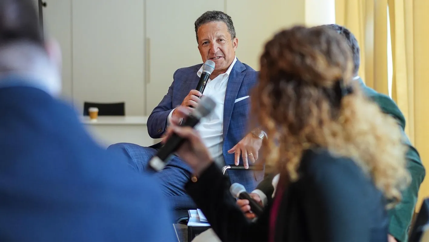A man in a blue suit sit talking into a microphone at an education session. 