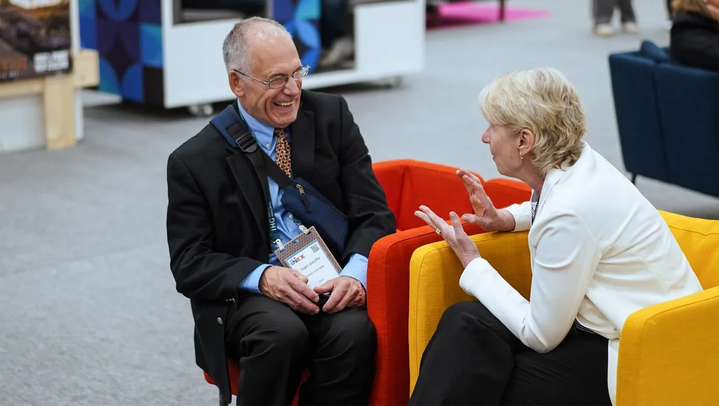 A man in a black suit sits laughing with a woman in a white jacket. They're sat on coloful chairs. 