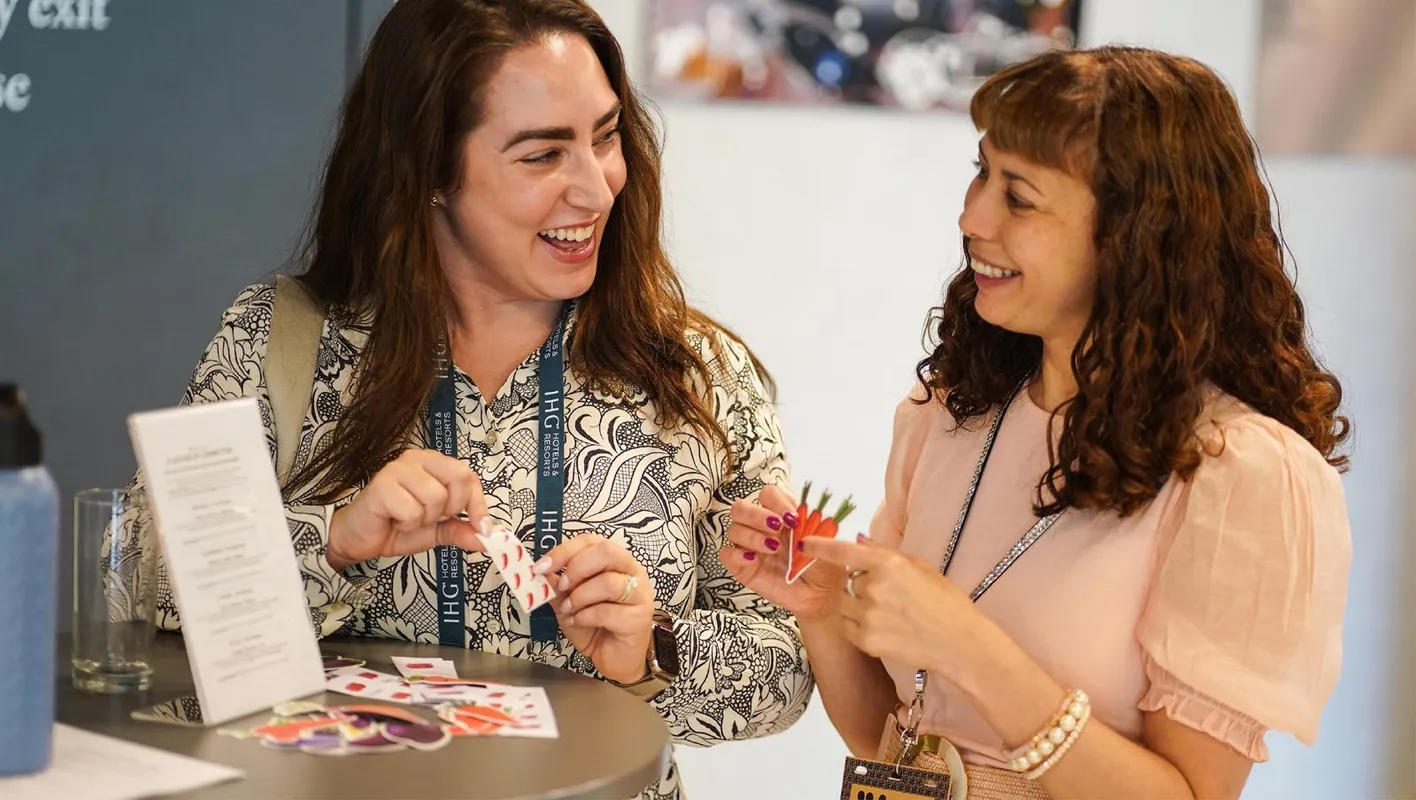 Two attendees in dresses stand smiling while interacting with stickers on a high table.