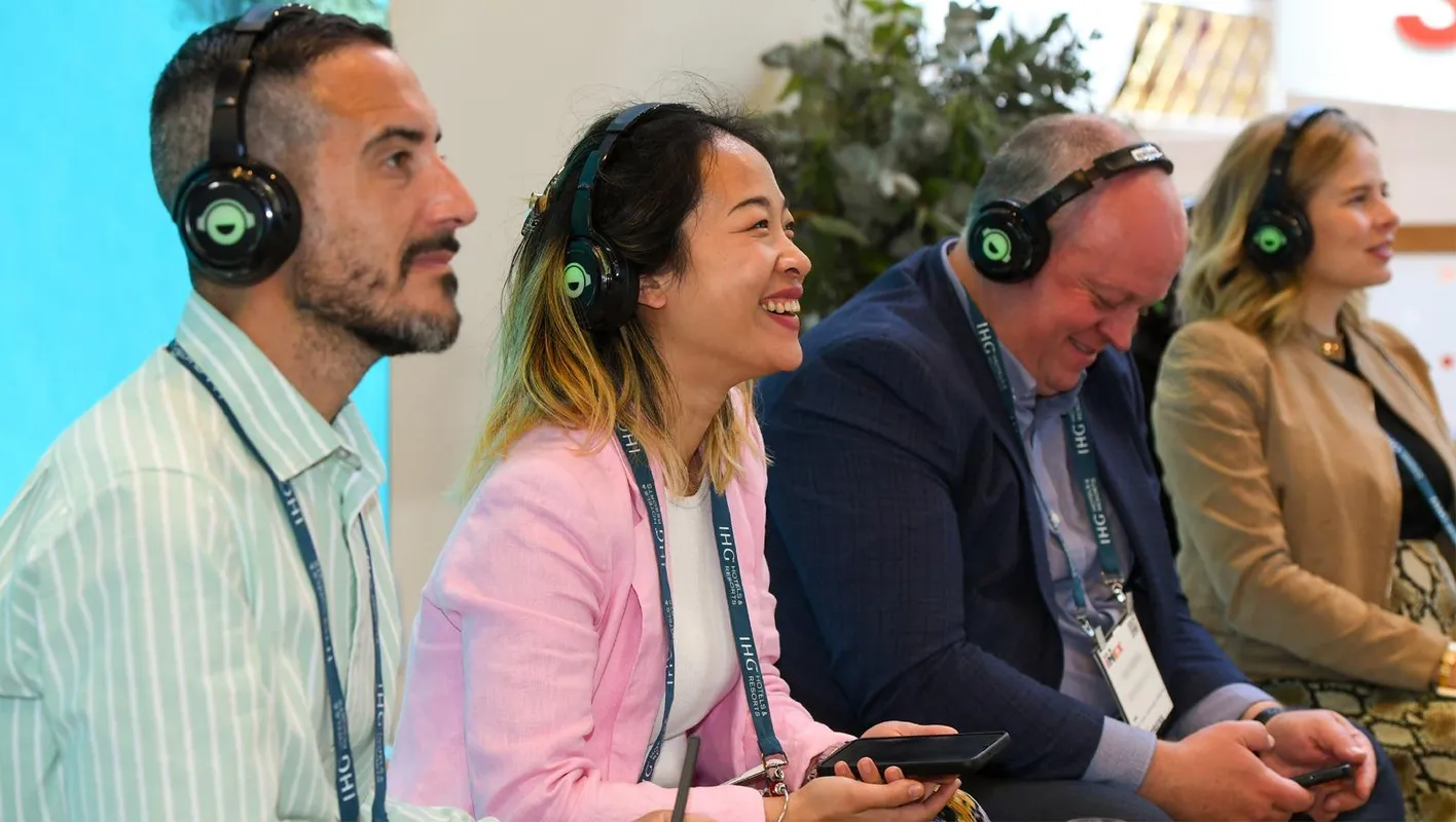 3 atttendees sit in a row laughing. They have headphones and landyards on, wearing business casual.