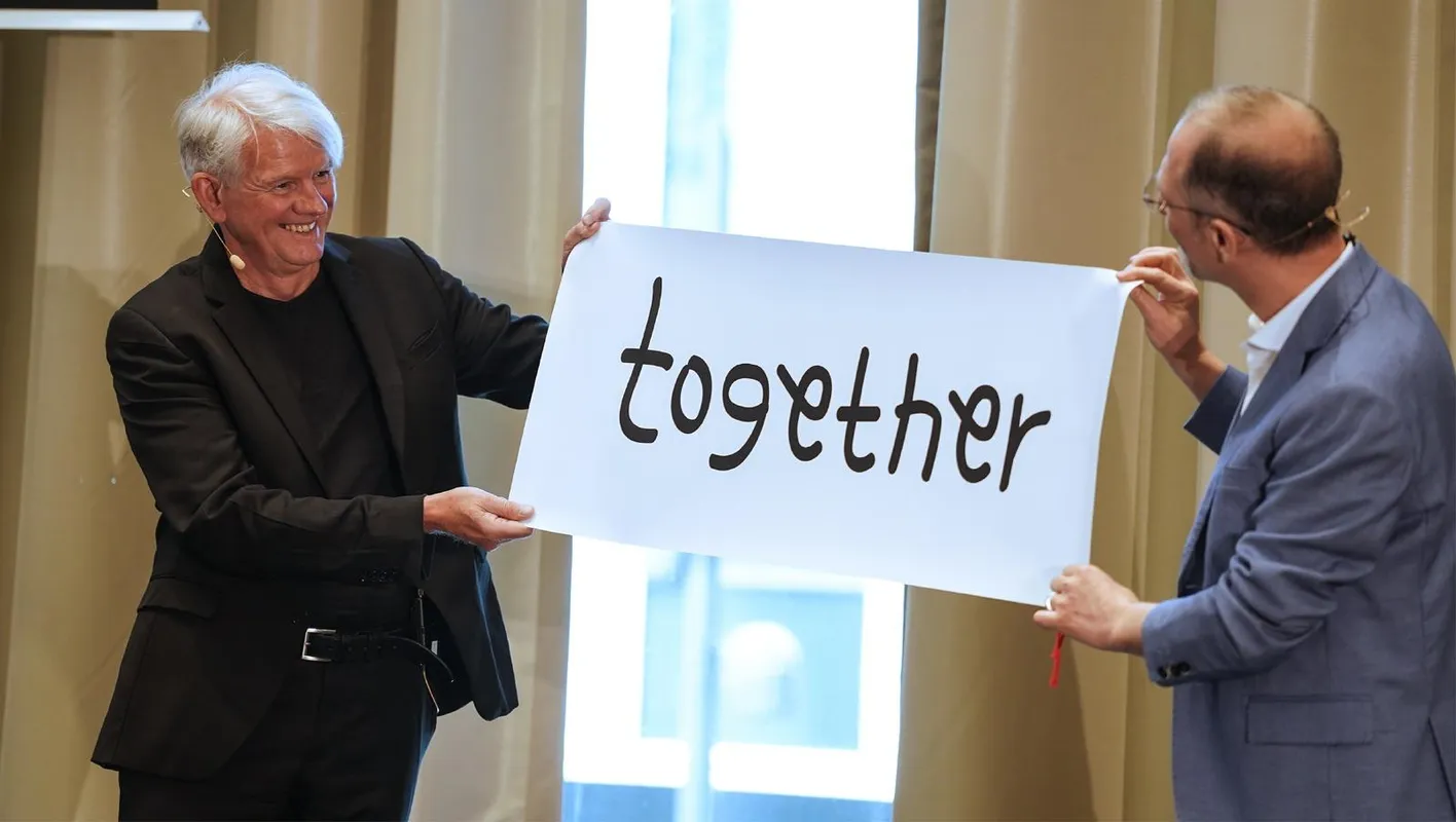 Two men in a confernce room hold a white sign that reads 'together. They're both wearing suits.