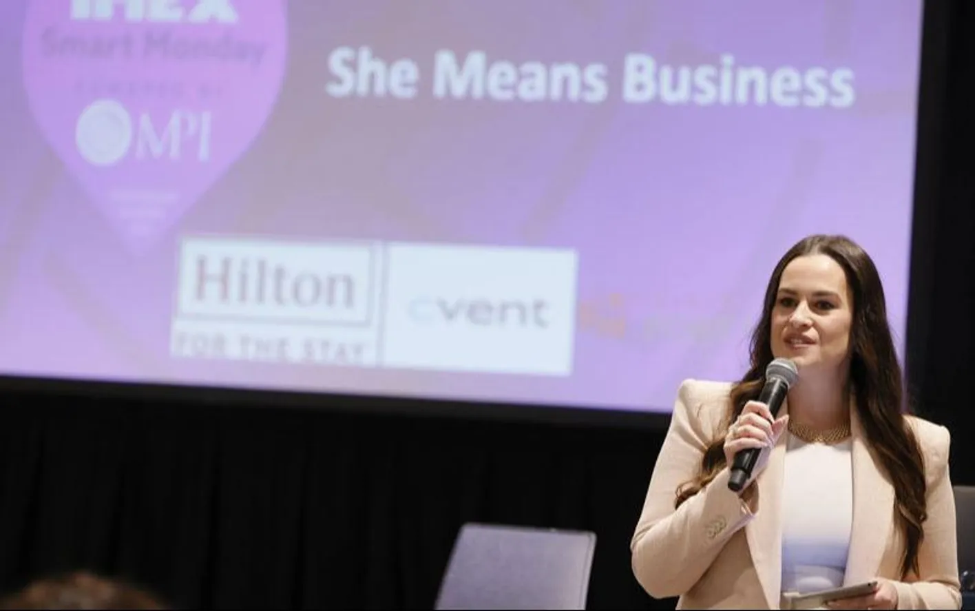  speaker presenting onstage with a microphone beneath a she means business backdrop