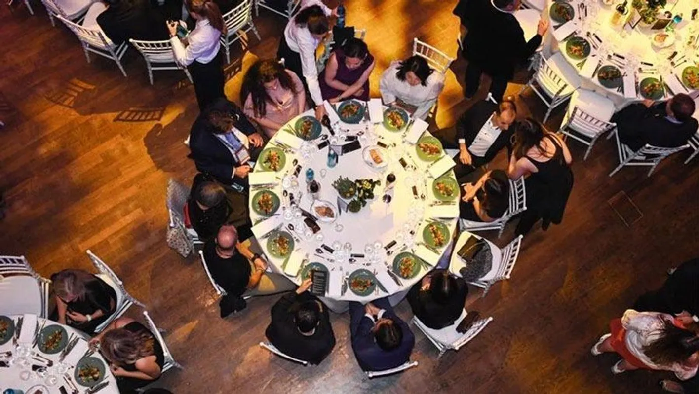 Aerial view of a round table of people talking and eating. There are green plates on the table.