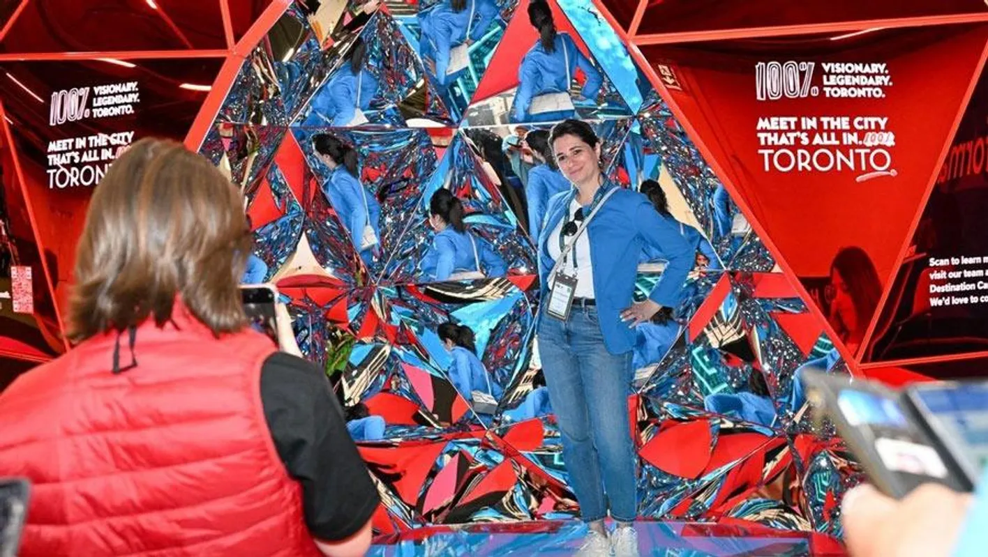 Attendee in a blue cardigan stands reflected in mirrored Toronto display as another takes a photo.