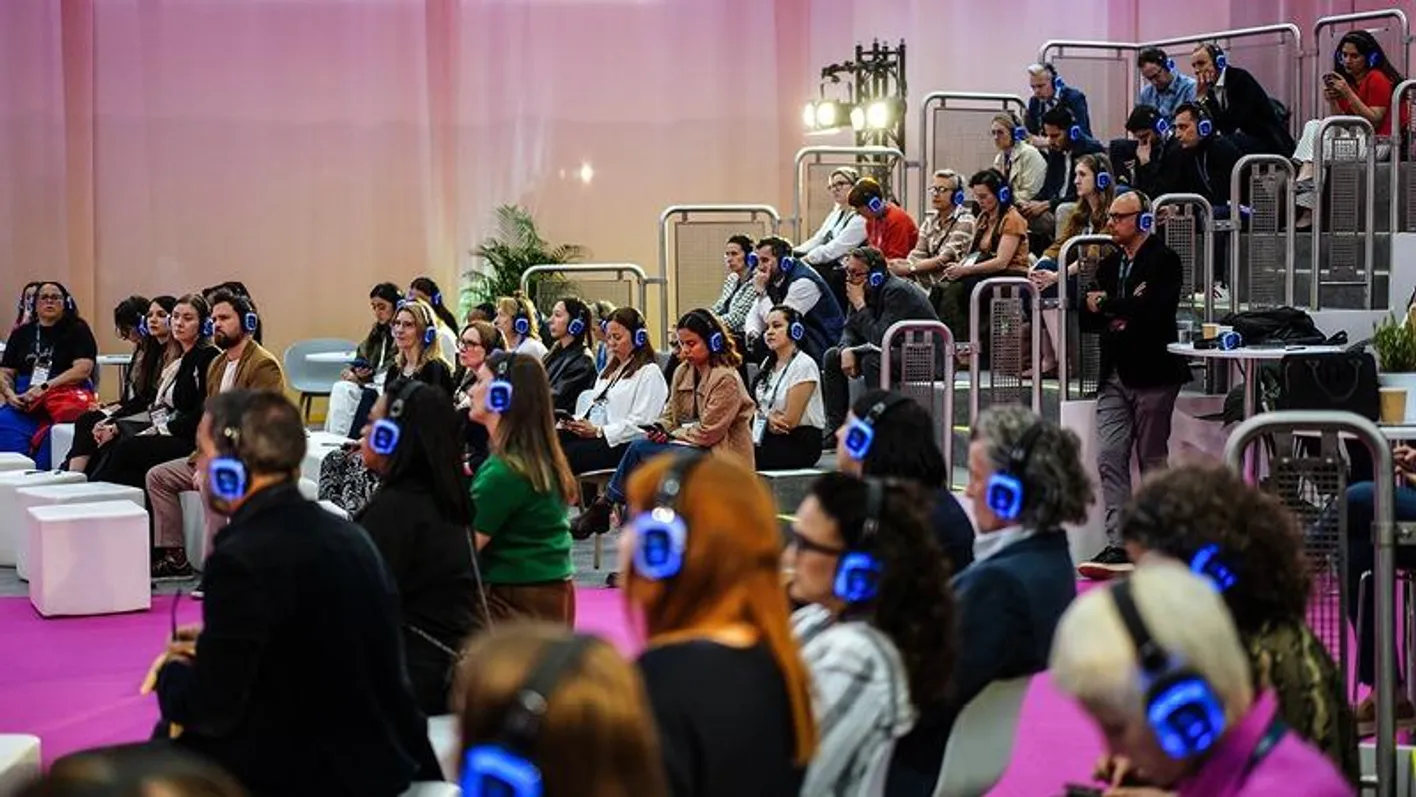 Large audience of attendees listen to an education session. They're wearing lanyards and headphones.