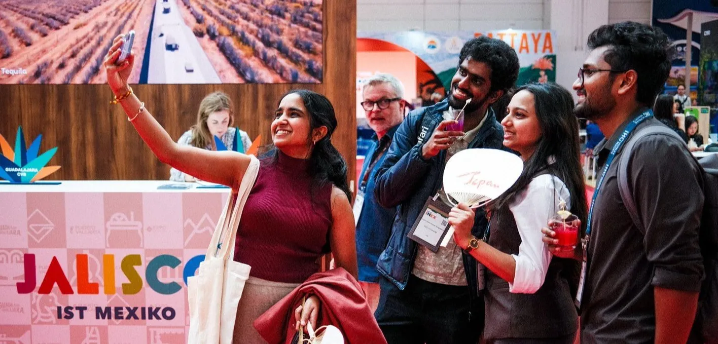 A group of people standing near an exhibitor booth taking a selfie, one person has a fan in her hand