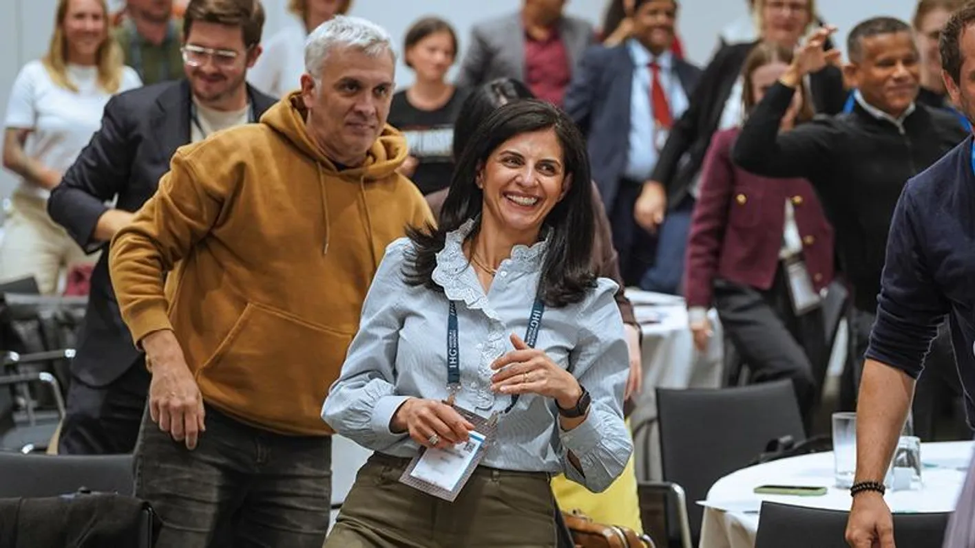 Two attendees stand smiling amongst a crowd, facing the stage. They're wearing business casual.