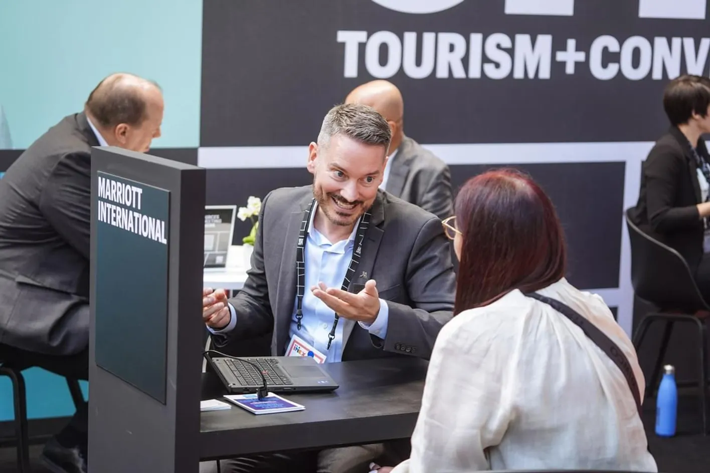 2 people in business casual clothing in a meeting on IMEX's show floorthey're in a dark grey booth.