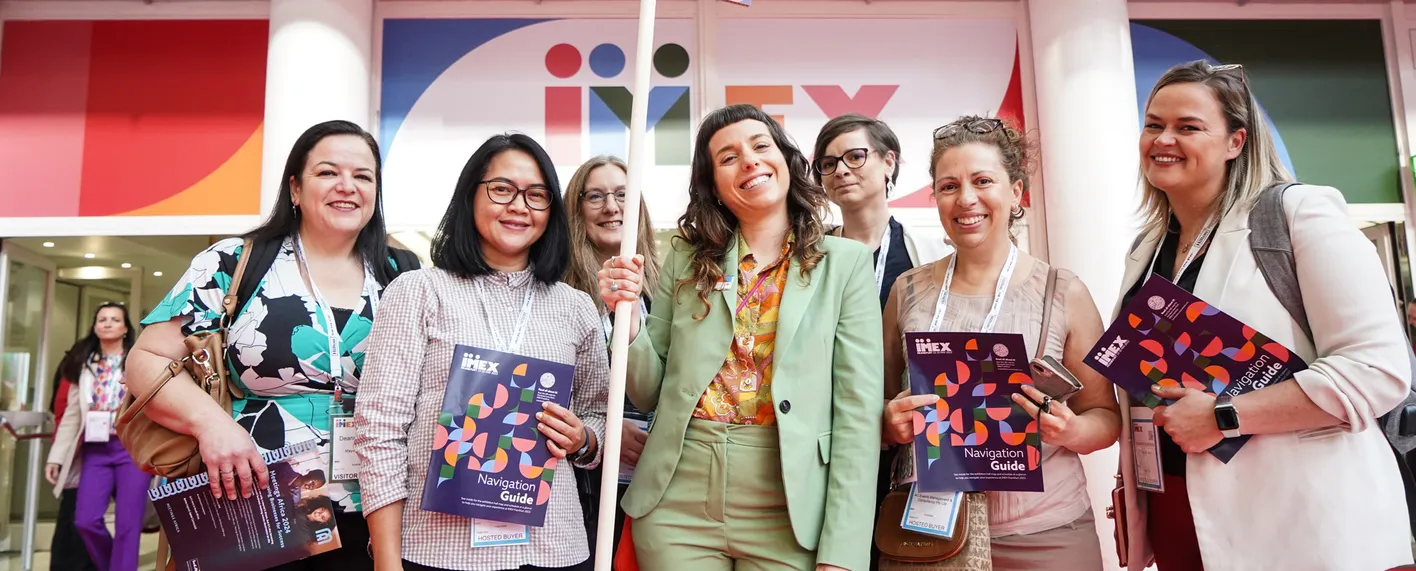 A group of attendees smiling in front of an IMEX banner. They're wearing business casual clothing.