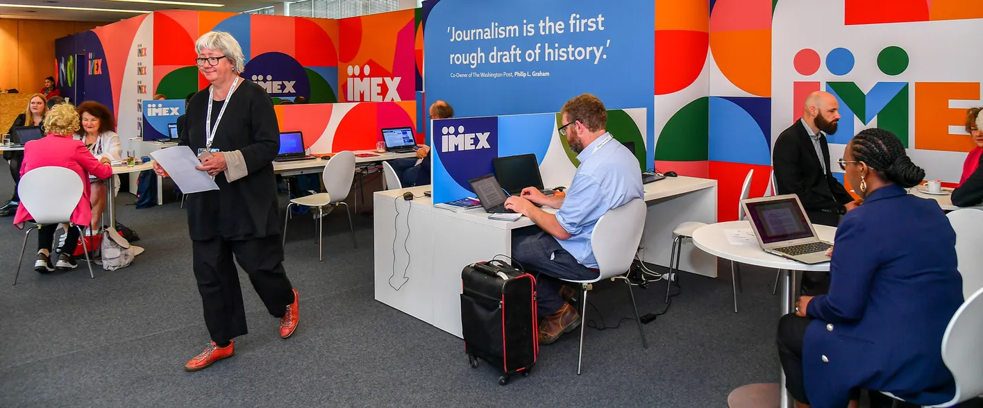 A colorful IMEX branded press centre containing members of the press working at their laptops.
