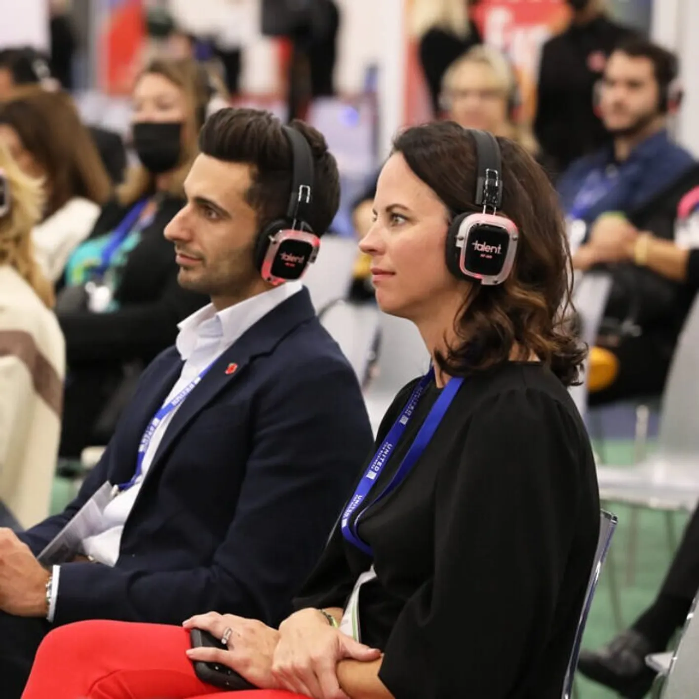 Two people seated at event wearing headphones labeled, other people are blurred in the background.