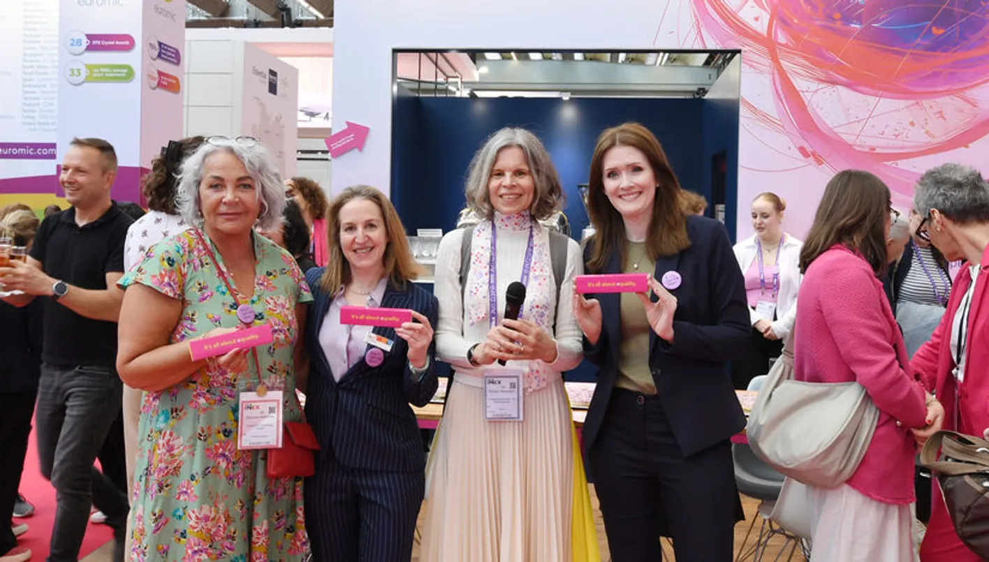 Five women stand holding pink name tags, with booths and banners in the background