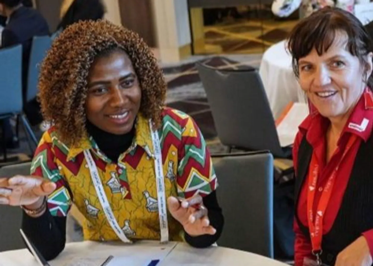 Two people seated indoors at a conference table, conversing, wearing lanyards and patterned clothing