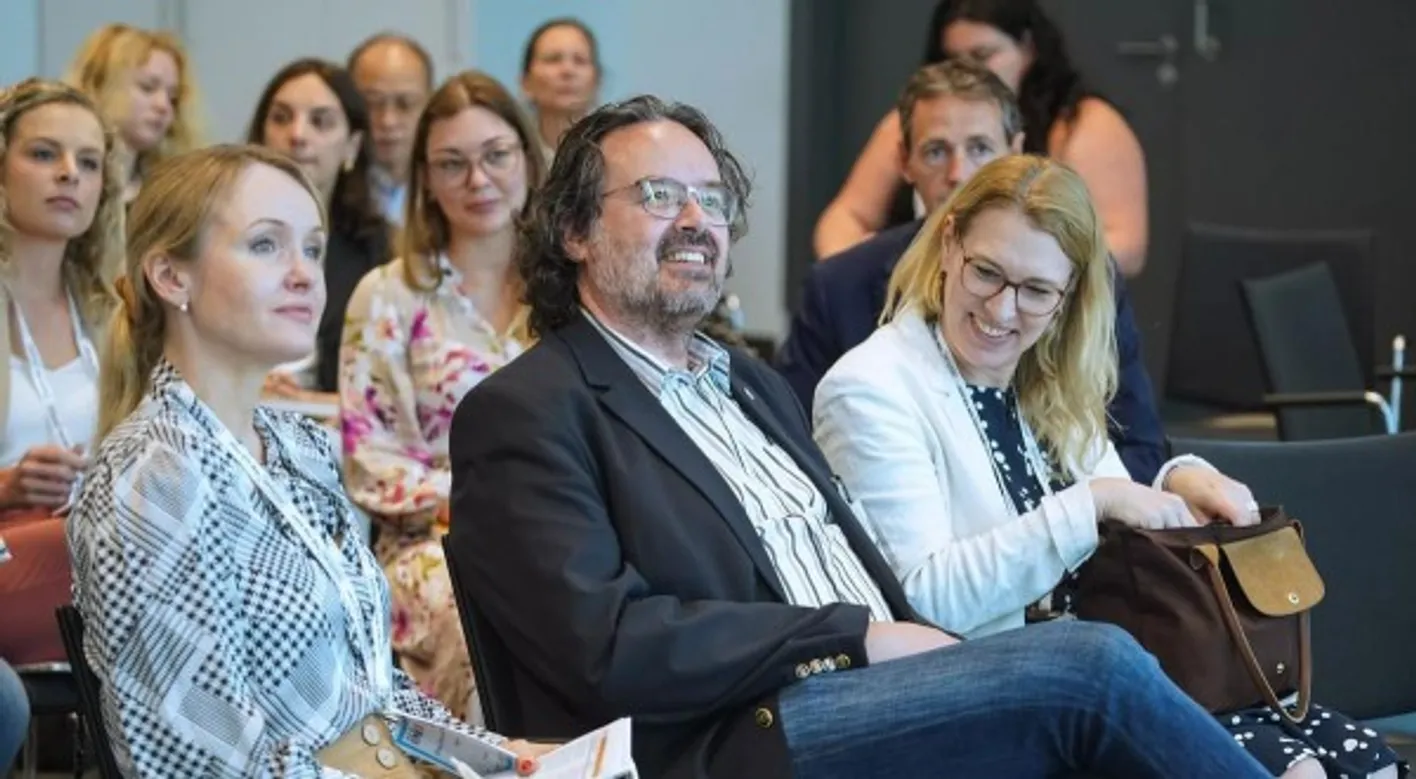 Attendees seated in a conference room, dressed in business casual, with notebooks and bags visible