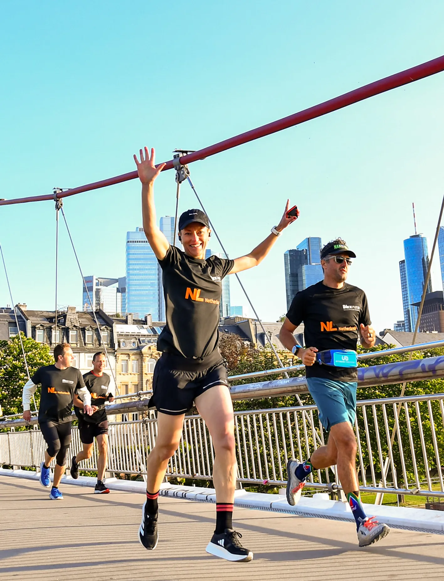 Runners in black 'NL' shirts on bridge with city skyline in background