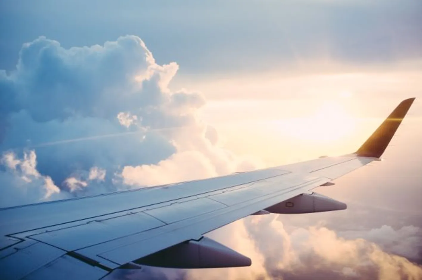 Image of a plane wing in flight in front of a cloudy sky with the sun peaking through the clouds.