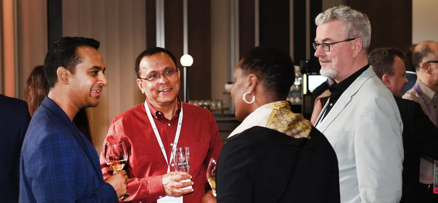 Four people conversing at a social event, holding wine glasses, with wooden panels behind them