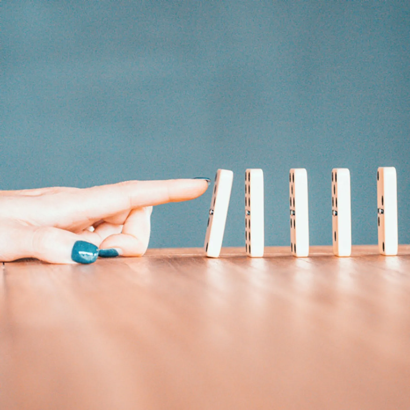 A finger points ready to push over a domino. There's five dominos in total all standing upright.