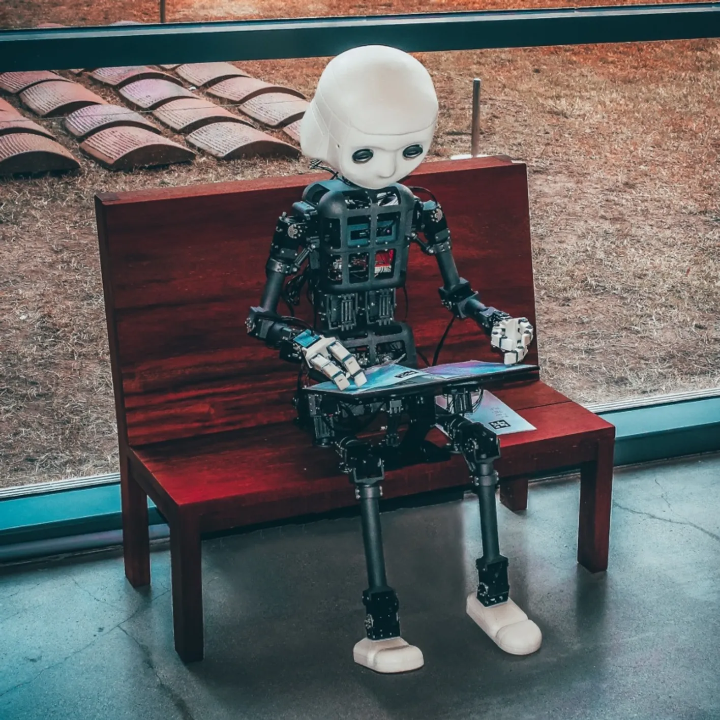 A robot sit on dark wood bench reading a magazine.