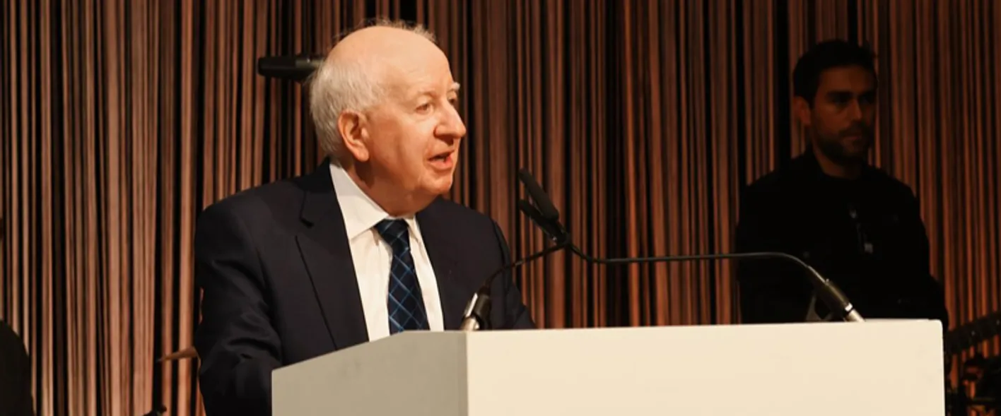 IMEX Chairman, Ray Bloom, talking at a plinth into a microphone in front of bronze colored curtains.