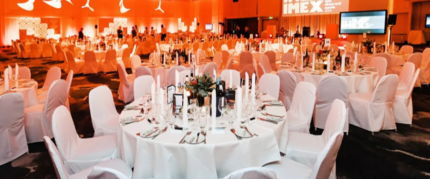 Several white roundtables set for a formal dinner. The chairs are also white. There's orange light