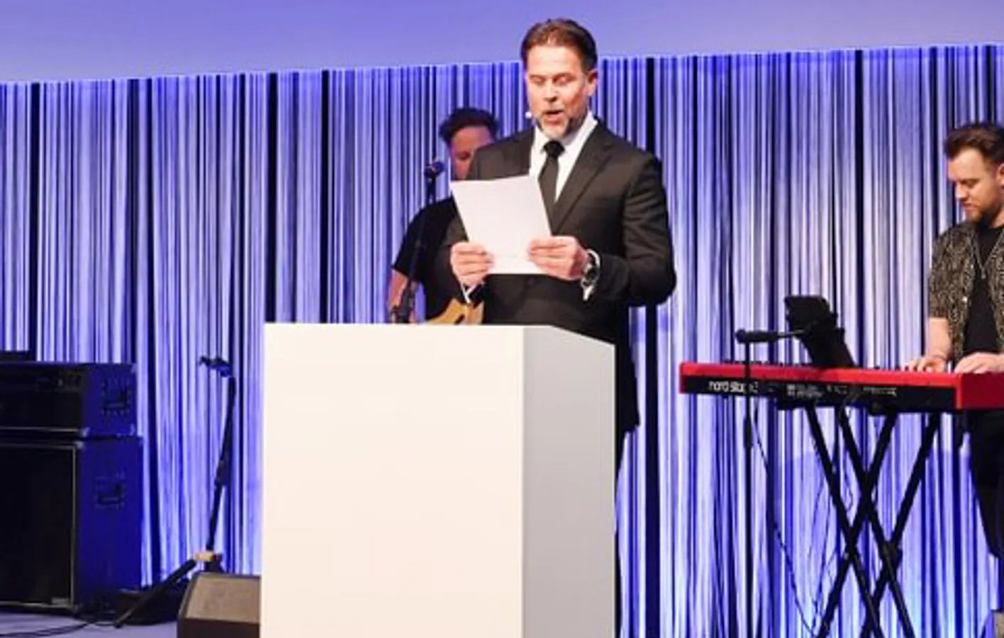 Gala Dinner MC stands at a plinth reading from a piece of paper. He's wearing a suit