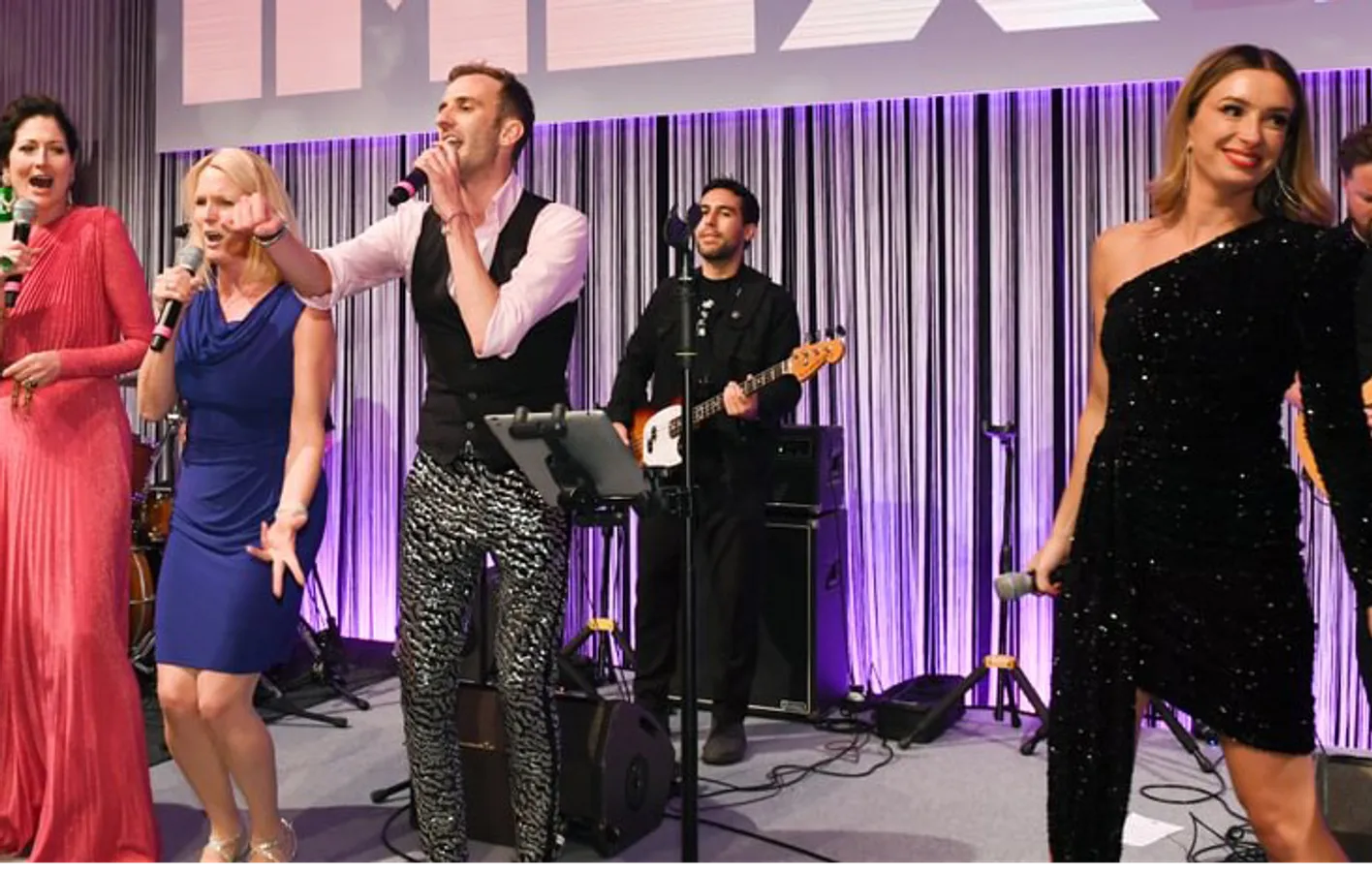 The band at the Gala Dinner stand on stage singing into microphones and playing instruments.