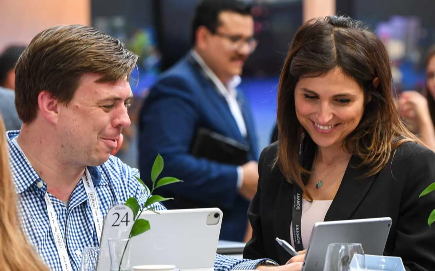 A man and a women, in business wear, looking at two tablets on a busy IMEX show floor. 
