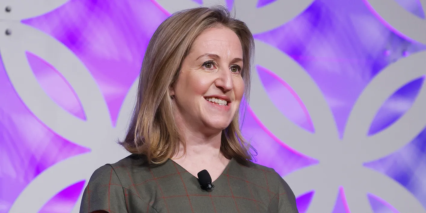 CEO of IMEX, Carina Bauer, stands looking beyond the camera smiling. She's wearing a checkered dress