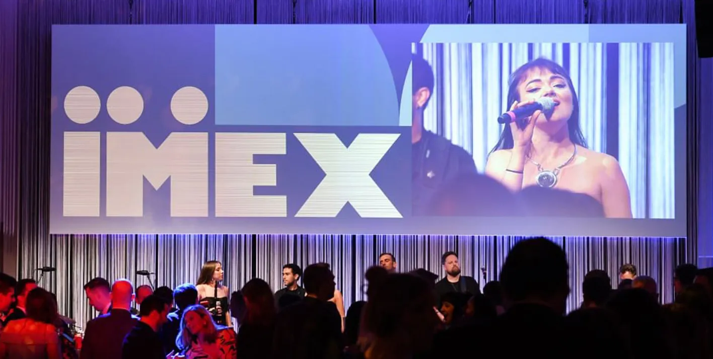 Crowd of Gala Dinner attendees in front of a large screen showing the IMEX logo and a woman singing