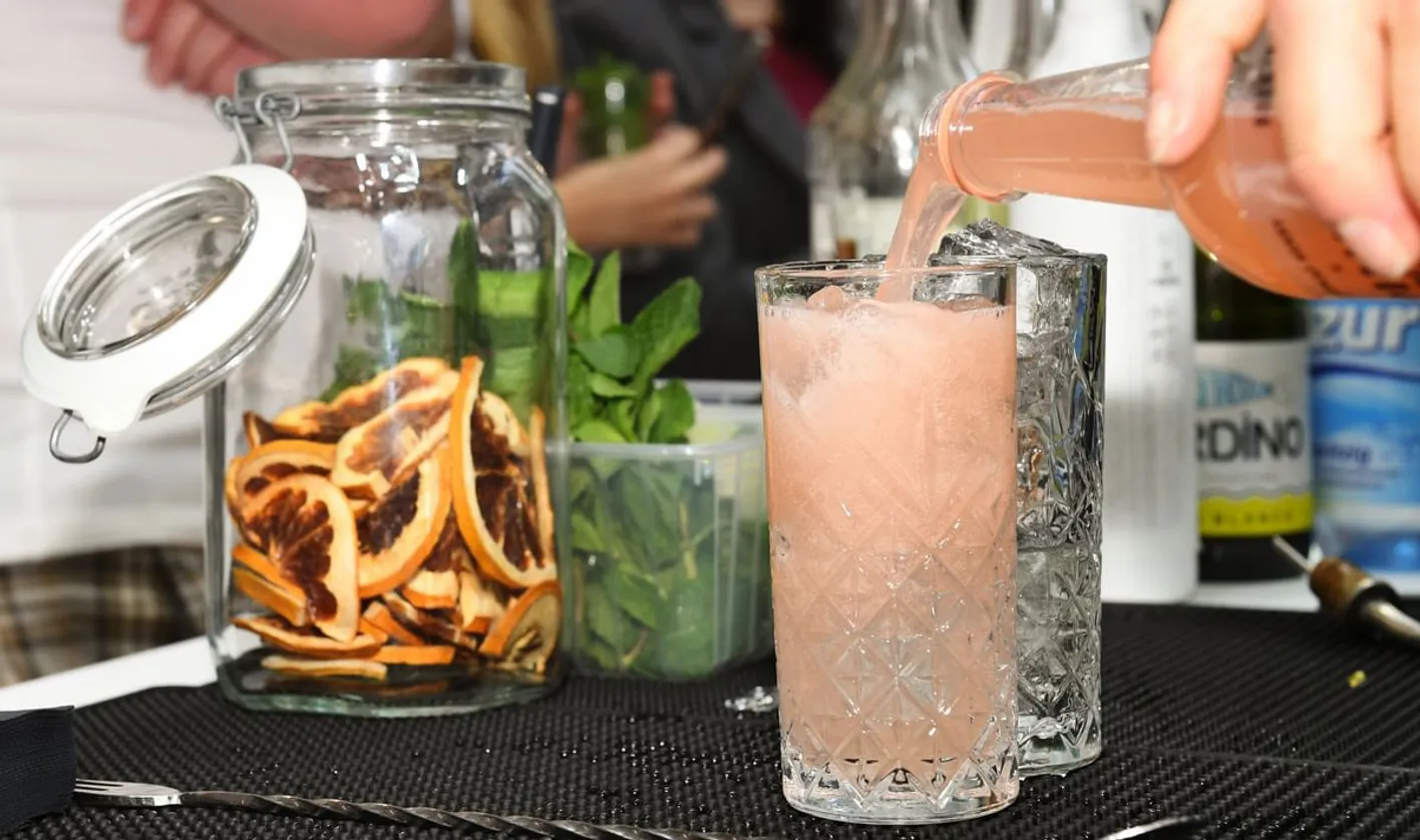 A light pink drink being poured into a glass. There's a jar of dried organges to the left.