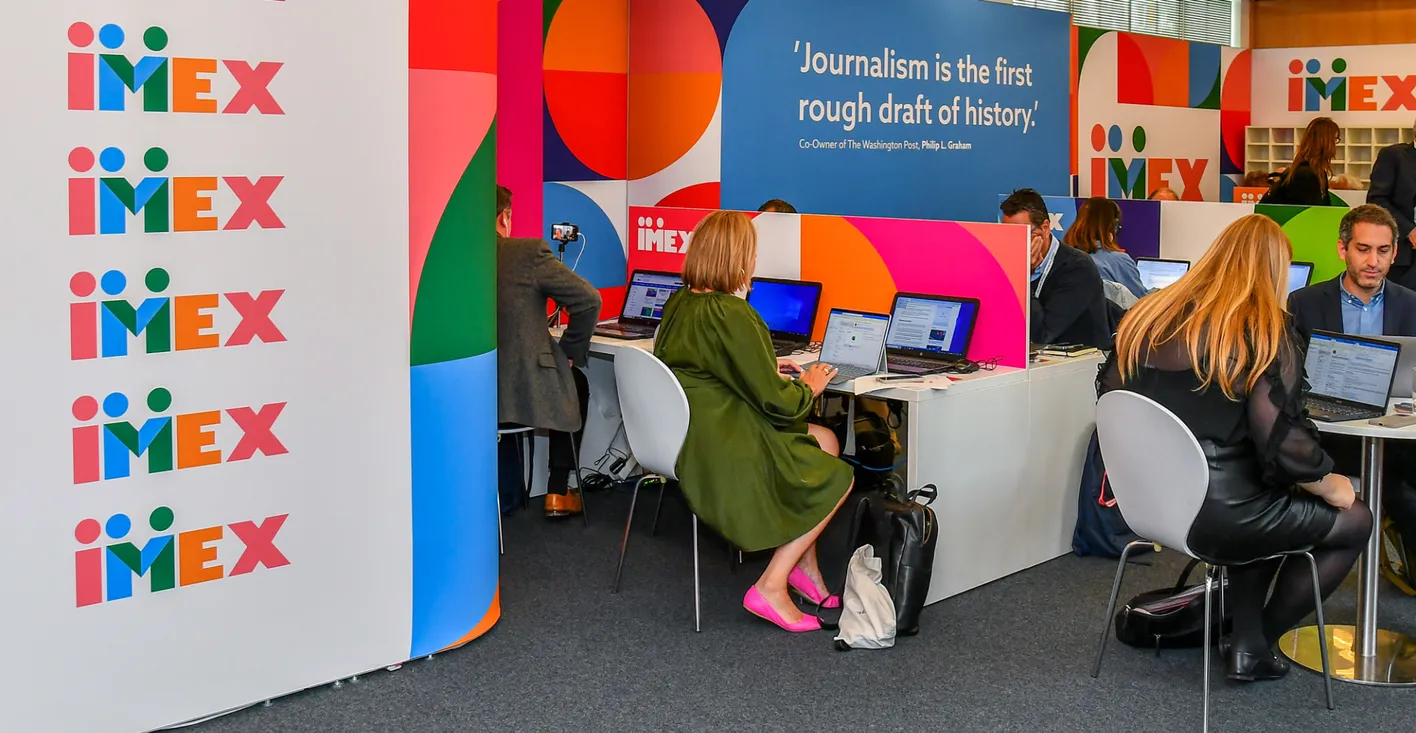 Press attendees all working on laptops with their backs to the camera. There's colorful signage