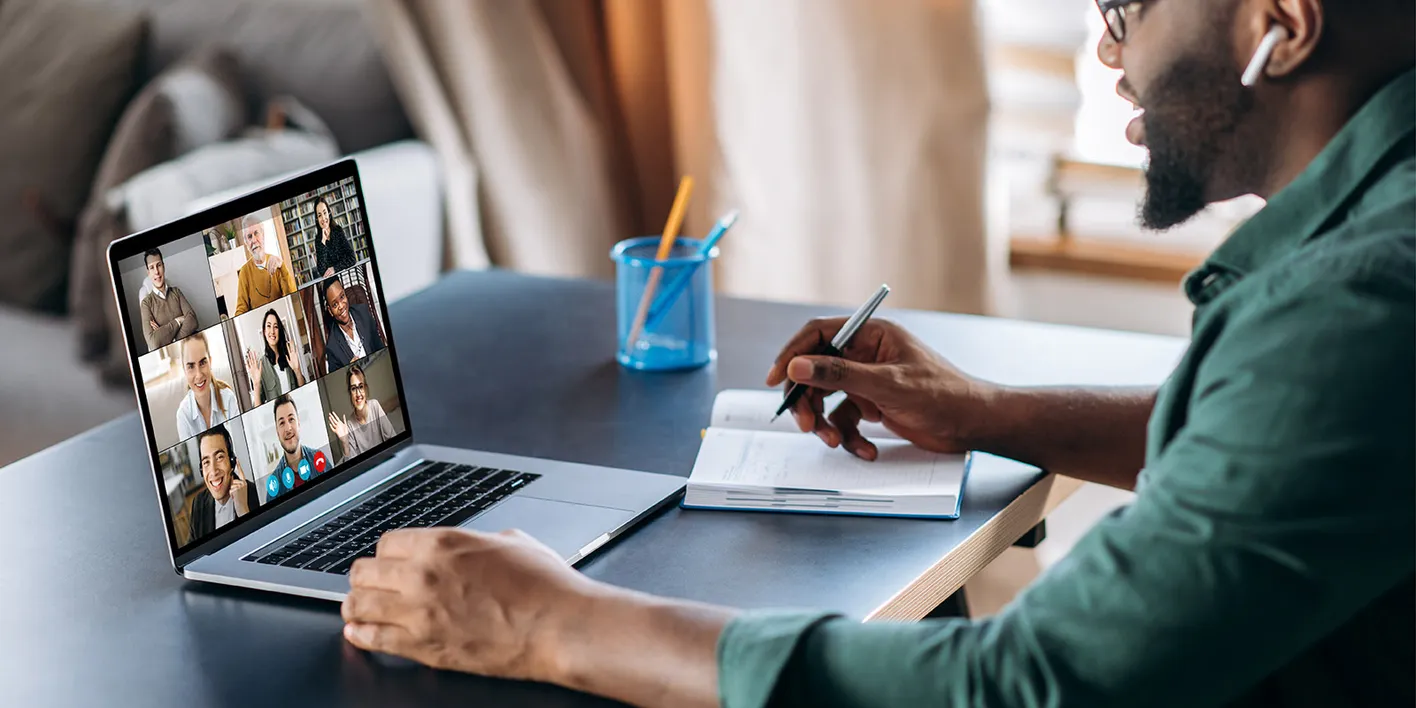 A person on a video call on a laptop, writing in notebook at desk with book and a pot of pens.