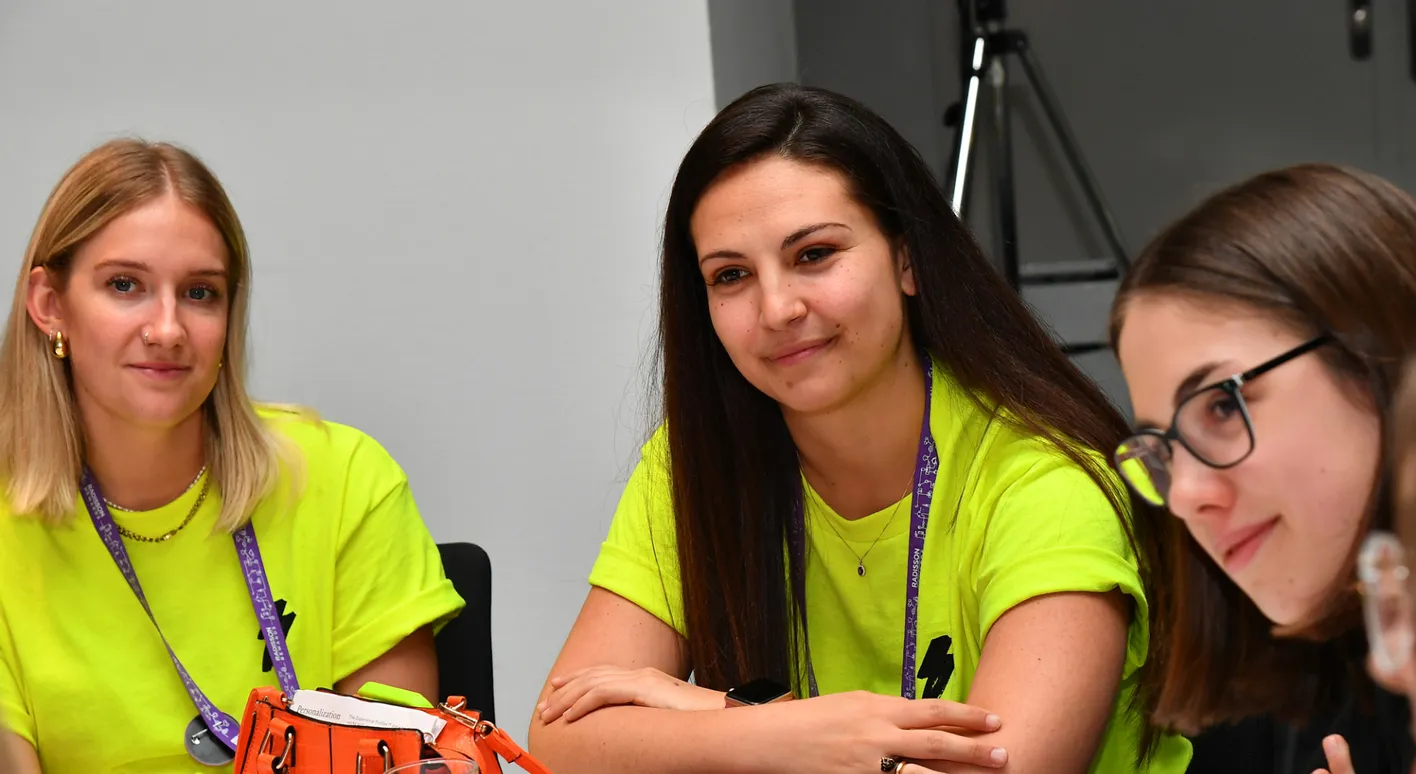 Two attendees in neon yellow shirts sit at a table smiling. They're wearing landyards.