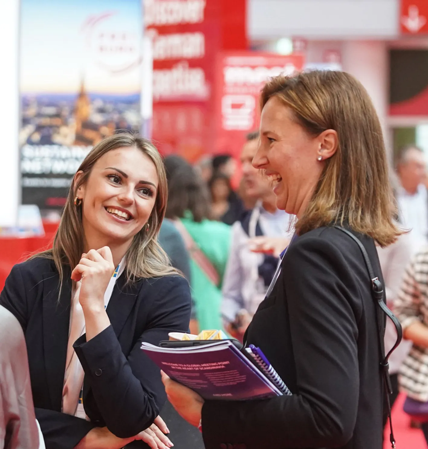 Two attendees stand on IMEX show floor laughing and smiling in business casual clothing.