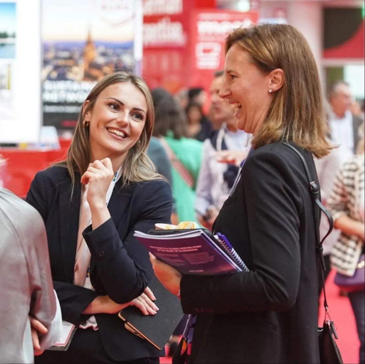 Two attendees stand on IMEX's show floor, laughing and smiling. They're wearing black blazers.