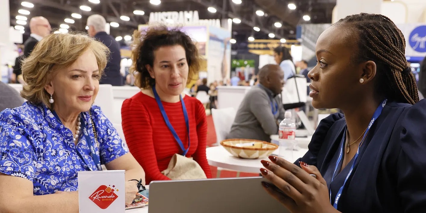 Two female buyers sitting with exhibitor at their booth and reviewing  information on a laptop