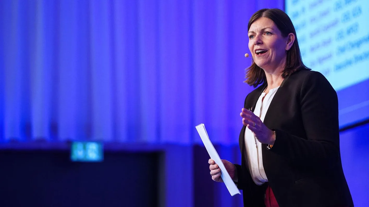 A woman holding a piece of page stands on a stage talking. She's wearing business casual.