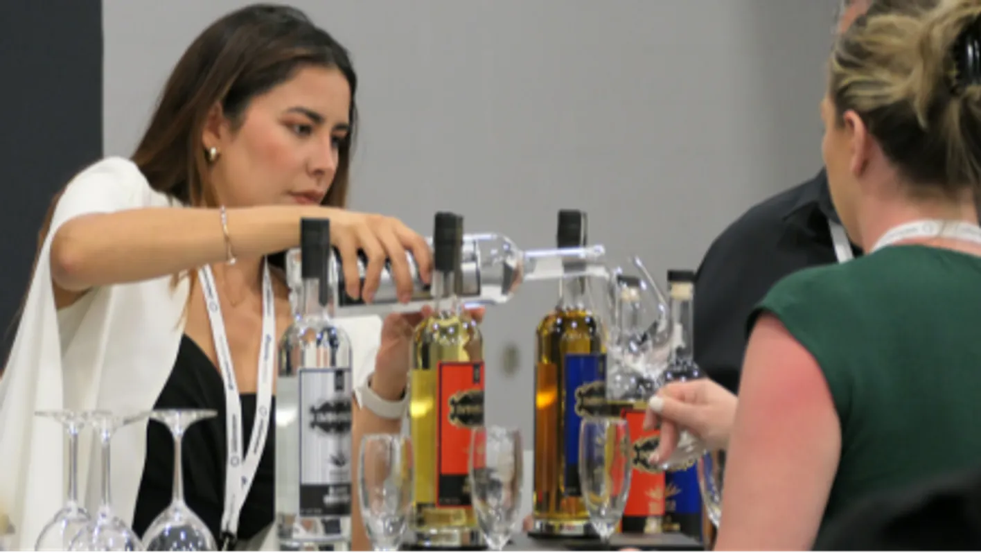 Two women, an exhibitor and a visitor. The exhibitor is pouring wine to the visitor's glass.